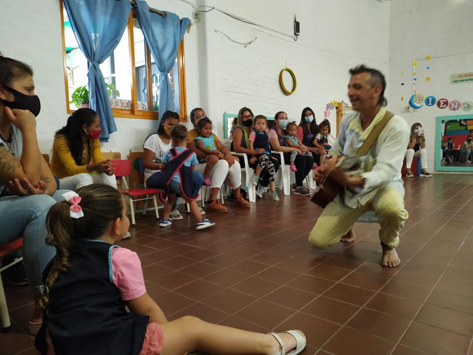 Mujeres, niñas y niños sentados vivencian la música que un hombre realiza con su guitarra