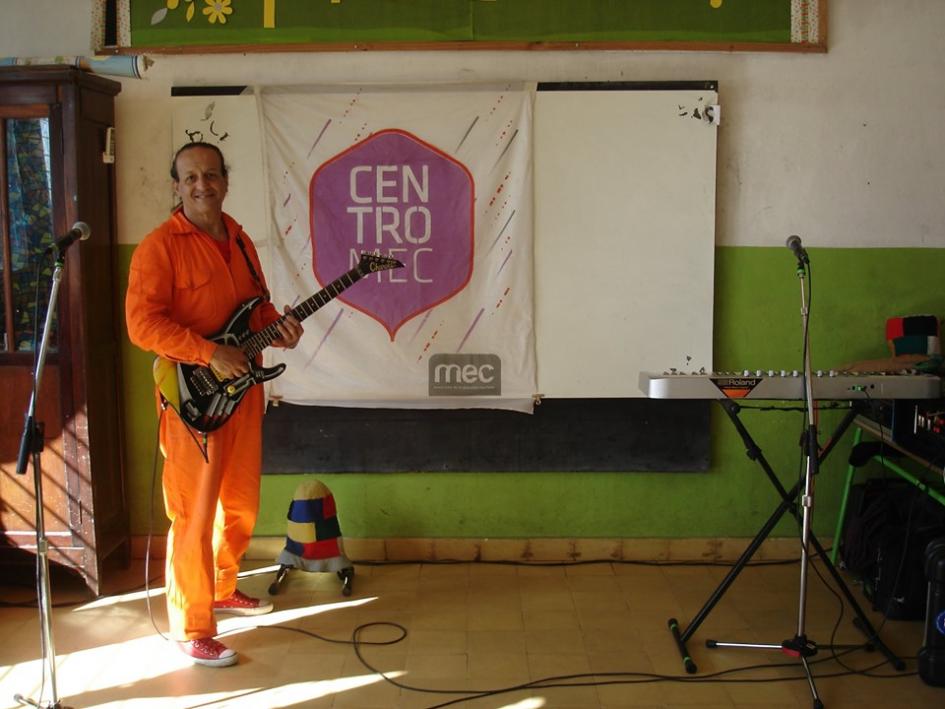 Integrante de Desolángeles con su guitarra. De fondo se encuentra la bandera de Centros MEC Integrante de Desolángeles con su guitarra. De fondo se encuentra la bandera de Centros MEC