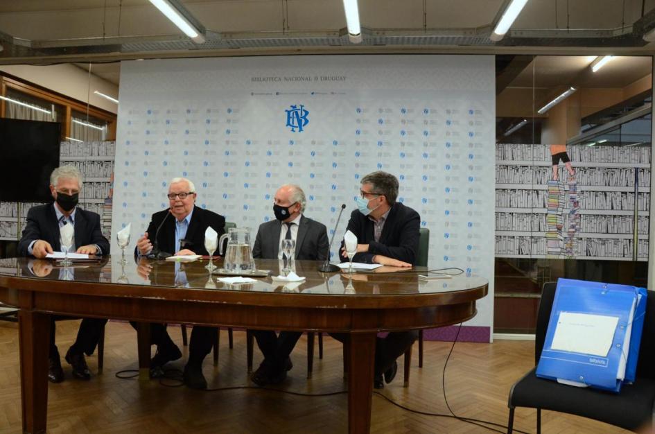 Autoridades sentadas y apoyan sus brazos en una mesa antigua y atrás el banner de la BNU