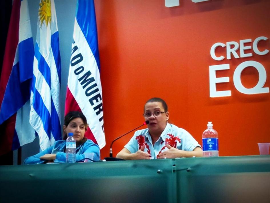 Con fondo rojo, banderas a la izquierda, hay dos personas sentadas, una de ellas hablando