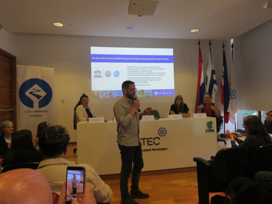 Encuentro de Rutas Unesco en Fray Bentos fortalece la promoción y gestión sostenible. Persona parada frente al público.