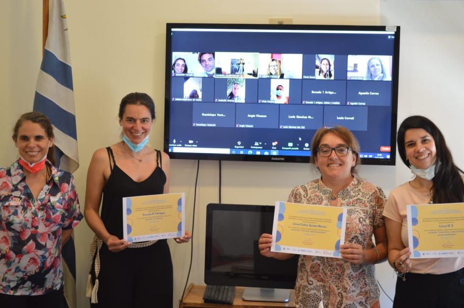 4 personas de pie, 3 de ellas mostrando el certificado de premiación. De fondo participante por zoom