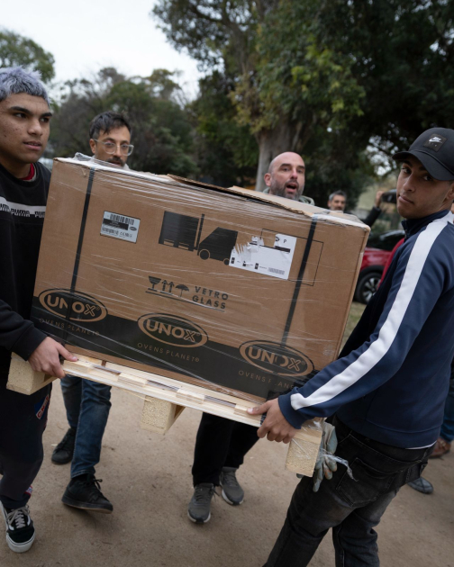 Taller de panadería de Cecap Brazo Oriental recibió horno prometido por Orsi Personas cargando la caja con el horno