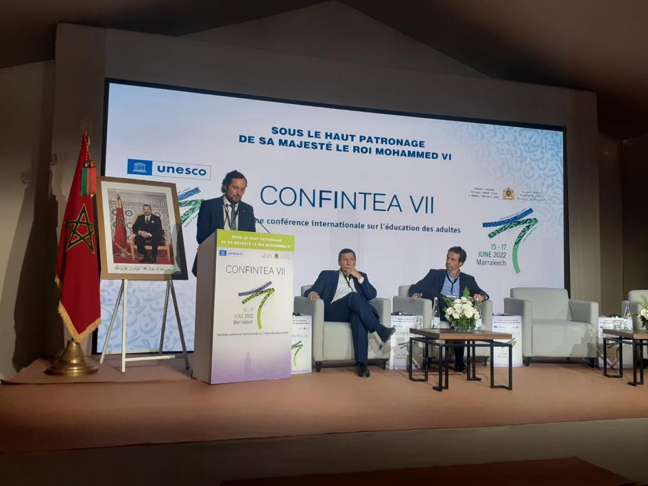 Exposición director Nacional de Educación, Gonzalo Baroni, en CONFINTEA VII. hombres sentado y hombre disertando en podio.