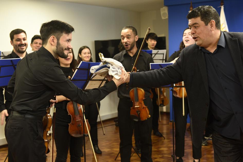 Un hombre dándole la bandera uruguaya a un músico. En el fondo, músicos con sus instrumentos