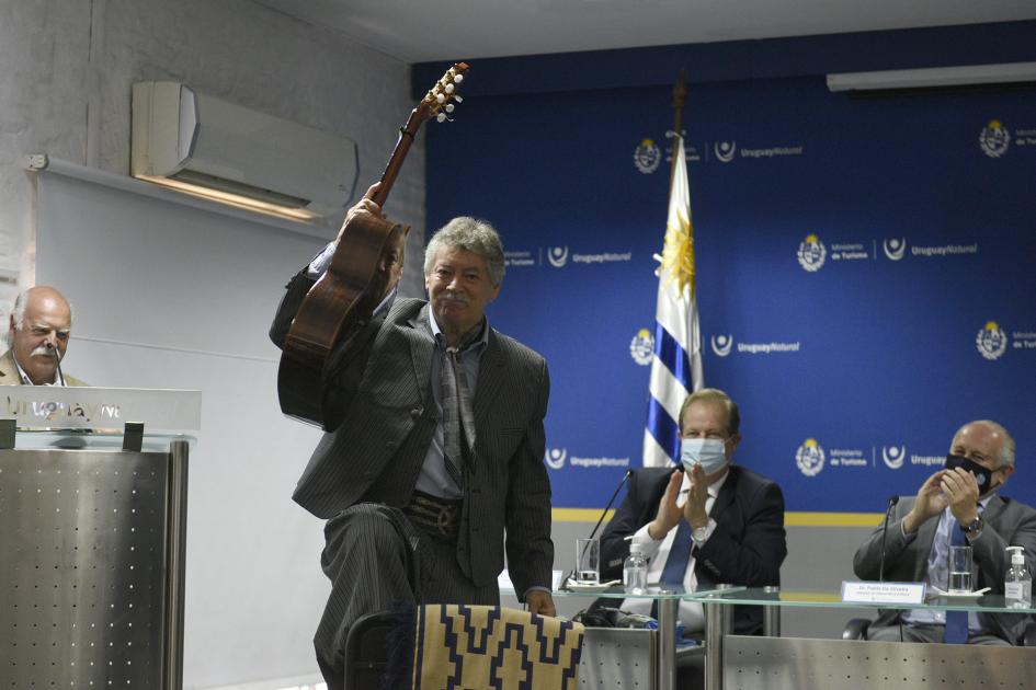 Gaucho parado con la guitarra en mano y atrás hay autoridades sentadas