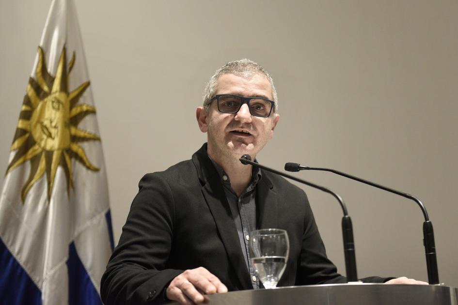 Hombre de lentes hablando y mirando la cámara. Atrás de ve la bandera uruguaya