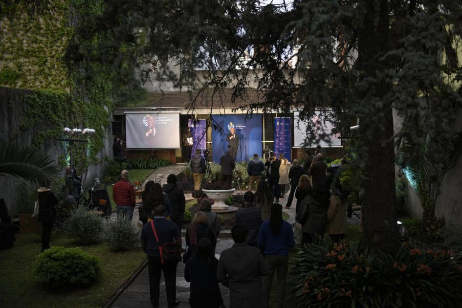 Plano general patio desde el público hacia el escenario Personas de pie y escenografía con logo MEC