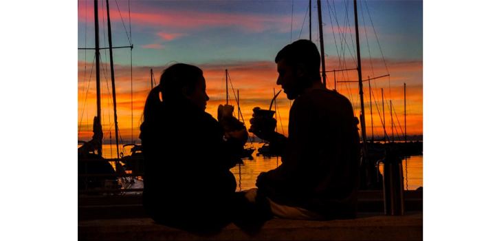 Dos personas tomando mate y comiendo tortas fritas con el atardecer de fondo