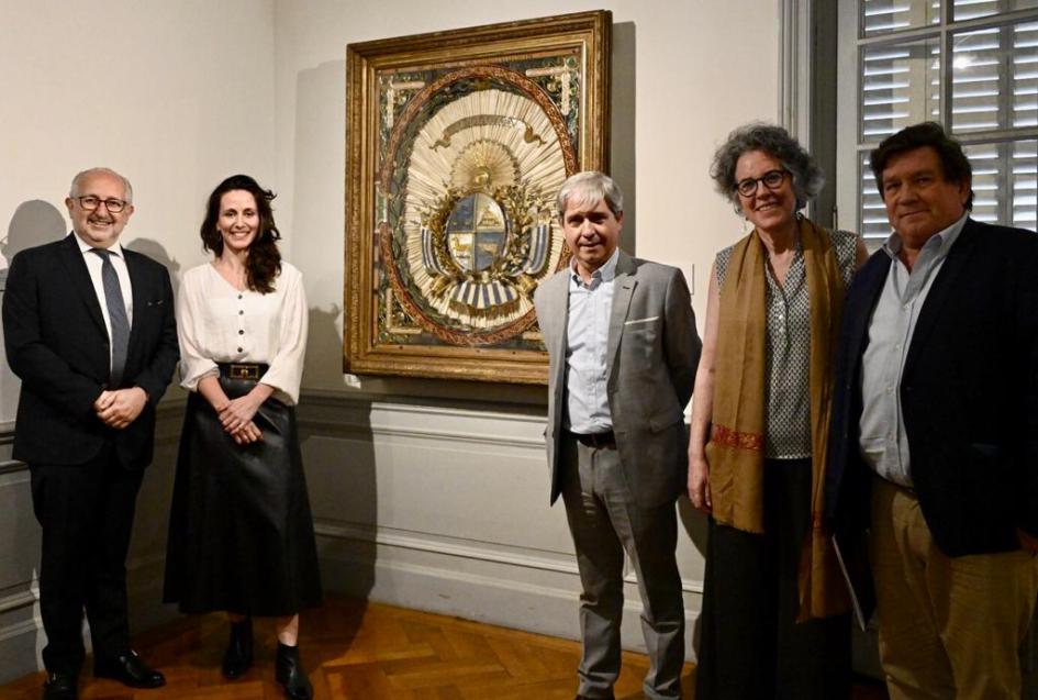 Autoridades en la ceremonia de apertura de la exposición. Personas paradas mirando de frente con cuadro de un escudo nacional al fondo.