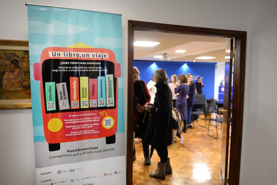 Lanzamiento campaña "Un libro, un viaje". En la puerta de la sala rollup con imagen de la campaña, en la sala participantes de la actividad.