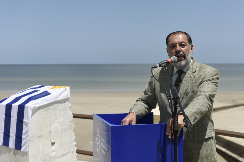 Hombre atrás del estrado dando su discurso. Atrás se ve la placa cubierta con la bandera uruguaya