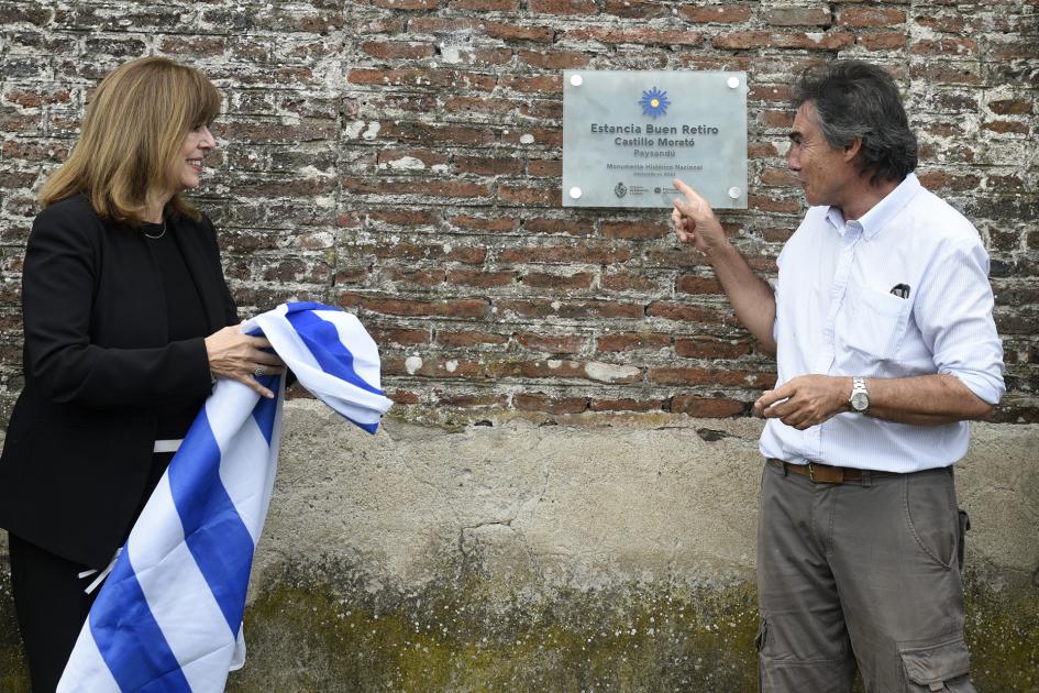 Mujer con la bandera uruguaya y hombre señalando la placa