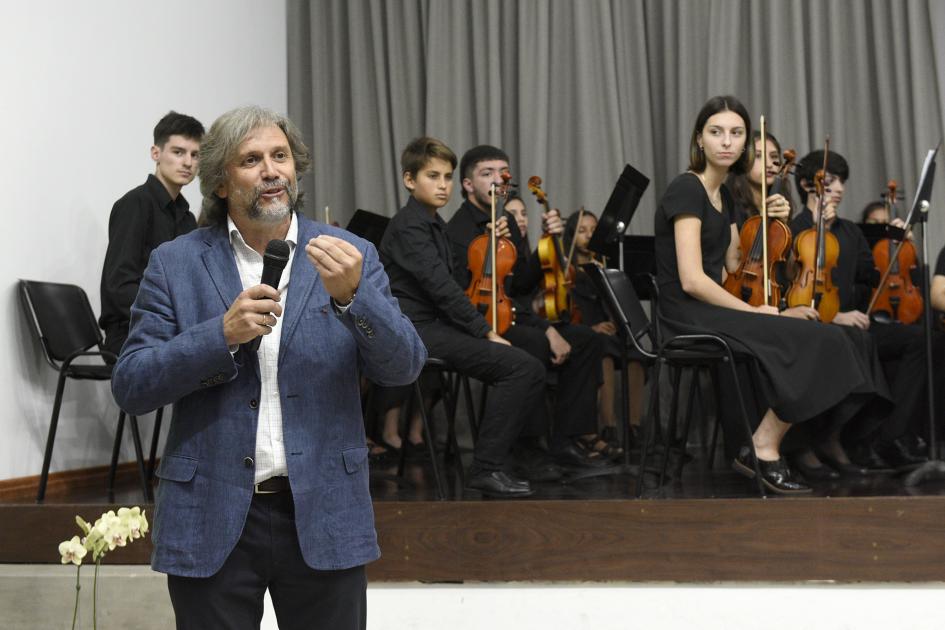 Hombre con micrófono en mano hablando y atrás se ve la Orquesta Juvenil del Sodre