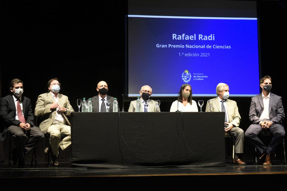 Jurado sentado en una mesa y Rafael Radi al medio. Atrás está la pantalla con el nombre del ganador
