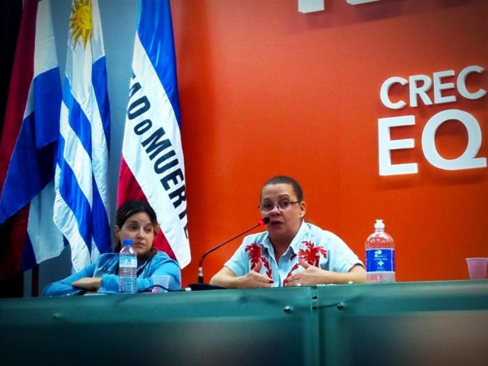 Con fondo rojo, banderas a la izquierda, hay dos personas sentadas, una de ellas hablando