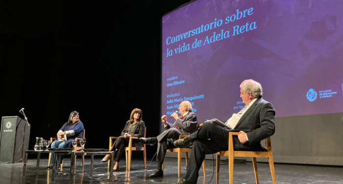 Participantes del conversatorio sentados en sillas en el escenario de la sala.