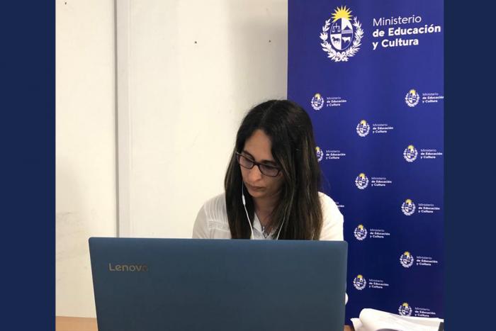 Se muestra una persona participando en una videoconferencia, con banner del MEC de fondo