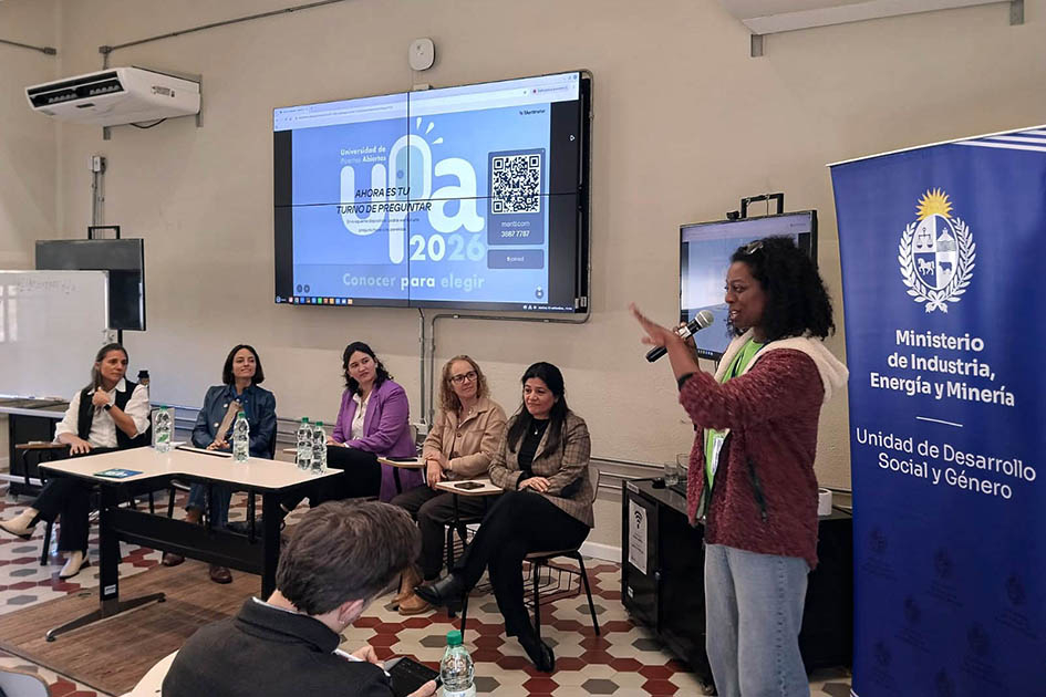 Panel Mujeres científicas en el sector público Panelistas, sentadas, mientras una mujer está de pie; al lado, banner; detrás, pantalla