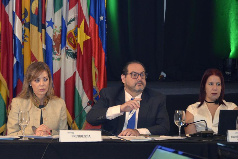 Un hombre señala, junto a dos mujeres, en la mesa de la Presidencia; detrás hay banderas