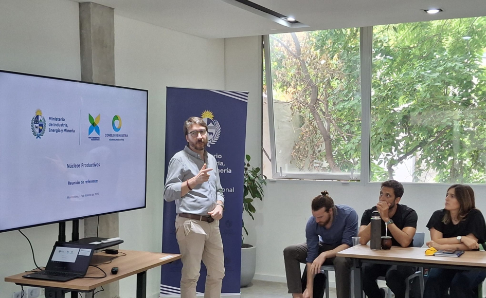 Míguez habla, de pie, ante personas sentadas en una mesa; hay ventanales y una pantalla