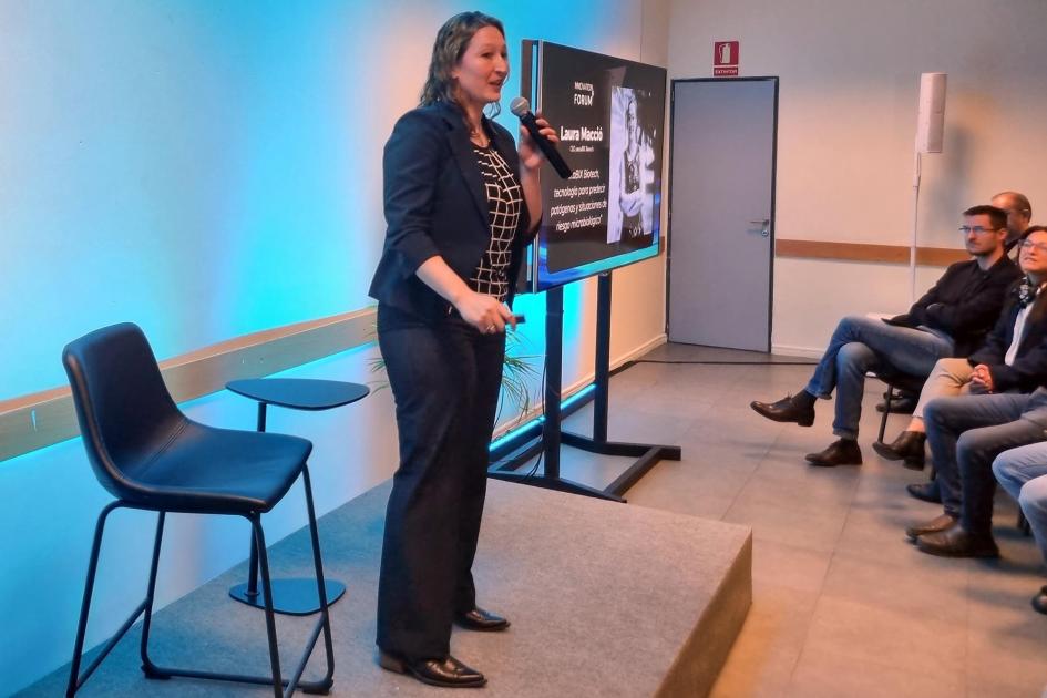 Laura Macció, de metaBIX Biotech Una mujer habla sobre un escenario; a sus lados, una silla y una pantalla; enfrente, el público