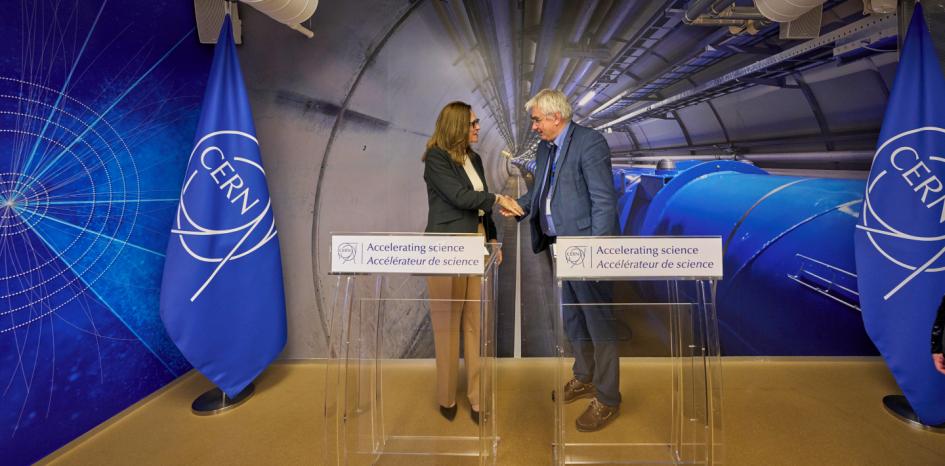 Ministra Elisa Facio y director Joachim Mnich Facio y Mnich se dan la mano, detrás de atriles; se ven banderas e imágenes del CERN