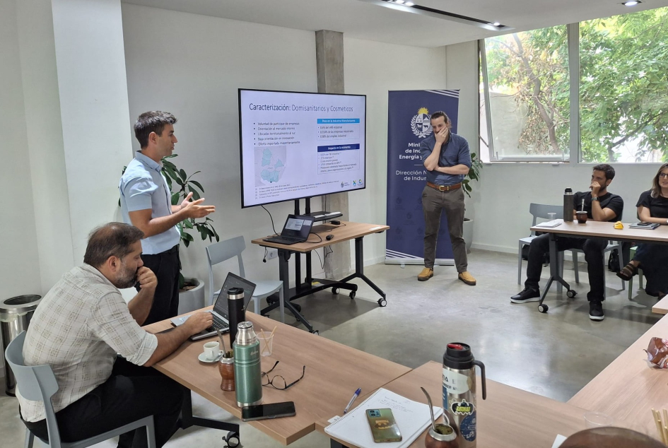 Personas de pie y sentadas en mesas, en un salón con ventanales y pantalla