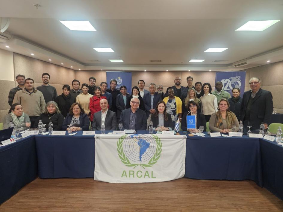 Foto grupal con participantes y expertos Grupo de asistentes al evento detrás de una mesa. Bandera de ARCAL visible.