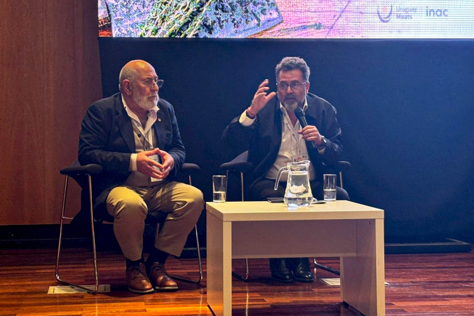 Carámbula habla, sentado, junto a Estévez; enfrente, mesa
