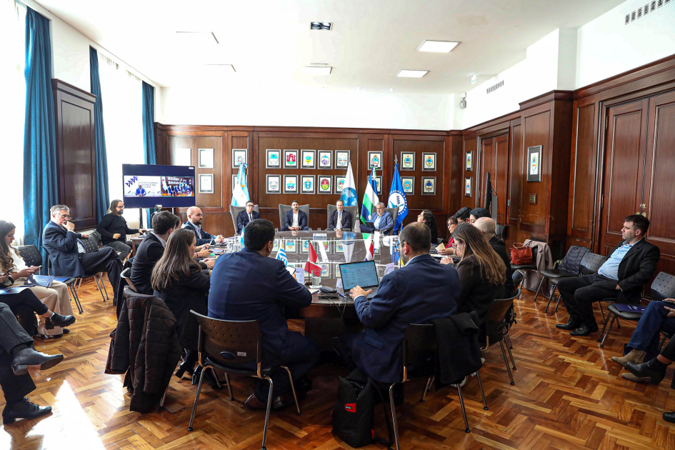 Personas reunidas alrededor de una mesa; contra la pared, en sillas, otras personas, sentadas