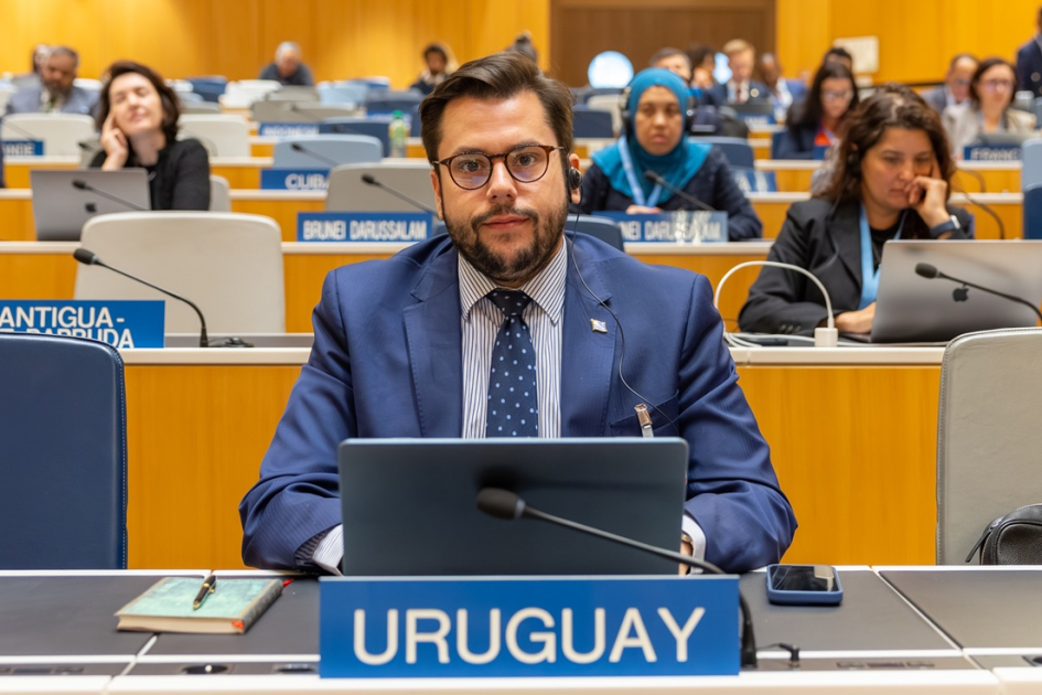 Persona sentada detrás de una mesa con un cartel de Uruguay; detrás, otras psersonas