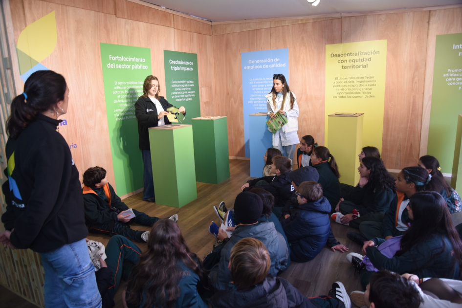  Cerca, estudiantes, sentados de espaldas; más lejos; encargadas del stand comparten información.