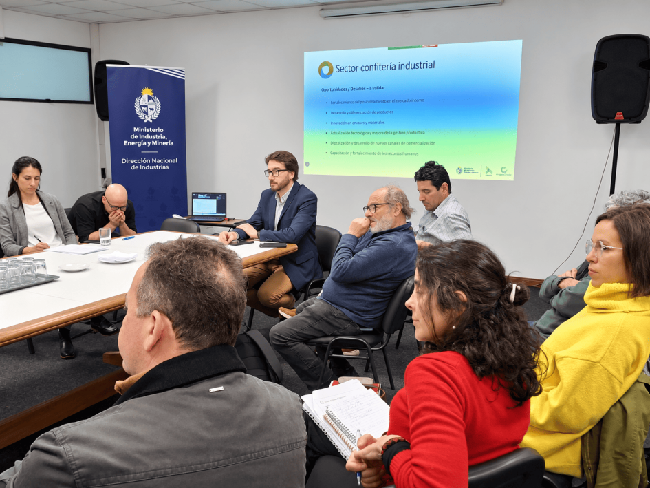 Autoridades presentes durante la reunión Míguez y Olesker, sentados en una mesa; frente a él, personas; al fondo, diapositiva en la pared