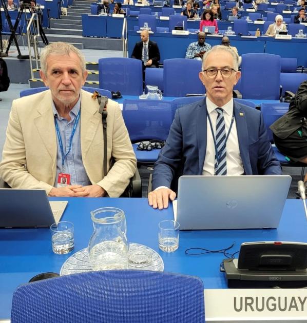 Director Gabriel González y embajador de Austria en Uruguay, Juan Carlos Ojeda Dos hombres están sentados en una mesa en la que hay una laptop; detrás se ven asientos azules