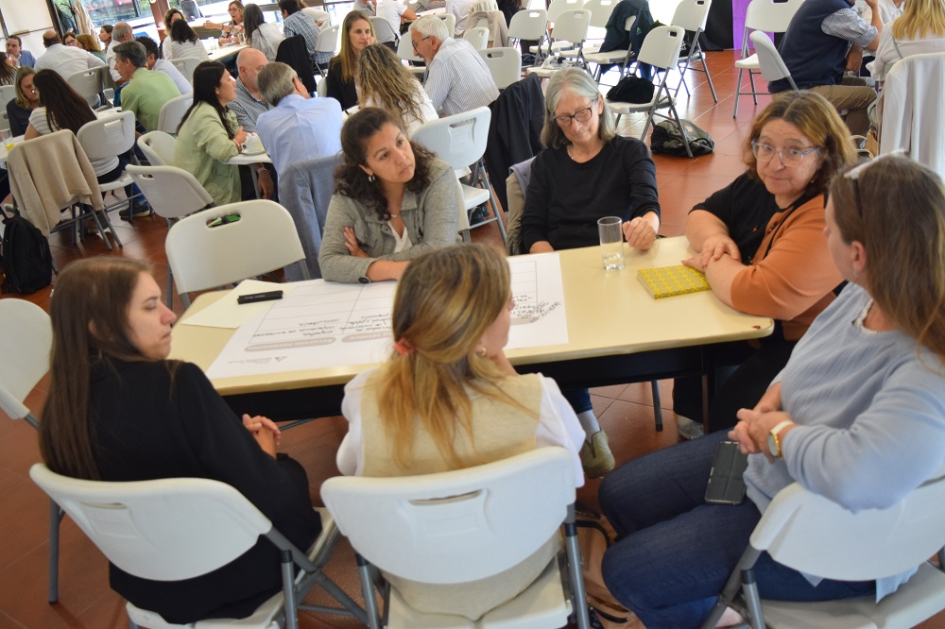 Personas sentadas alrededor de mesa. Detrás se ve más gente en mesas de trabajo