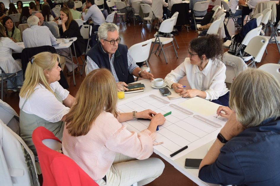 Personas sentadas alrededor de mesa. Detrás se ve más gente en mesas de trabajo