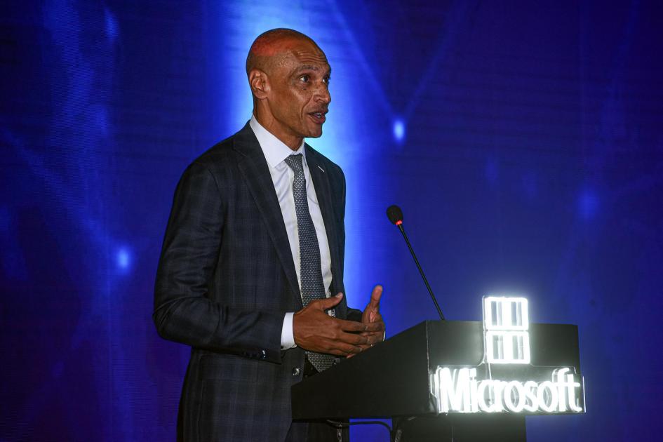 Vicepresidente ejecutivo de Microsoft, Christopher Young Young habla desde un atril; detrás, pantalla violeta