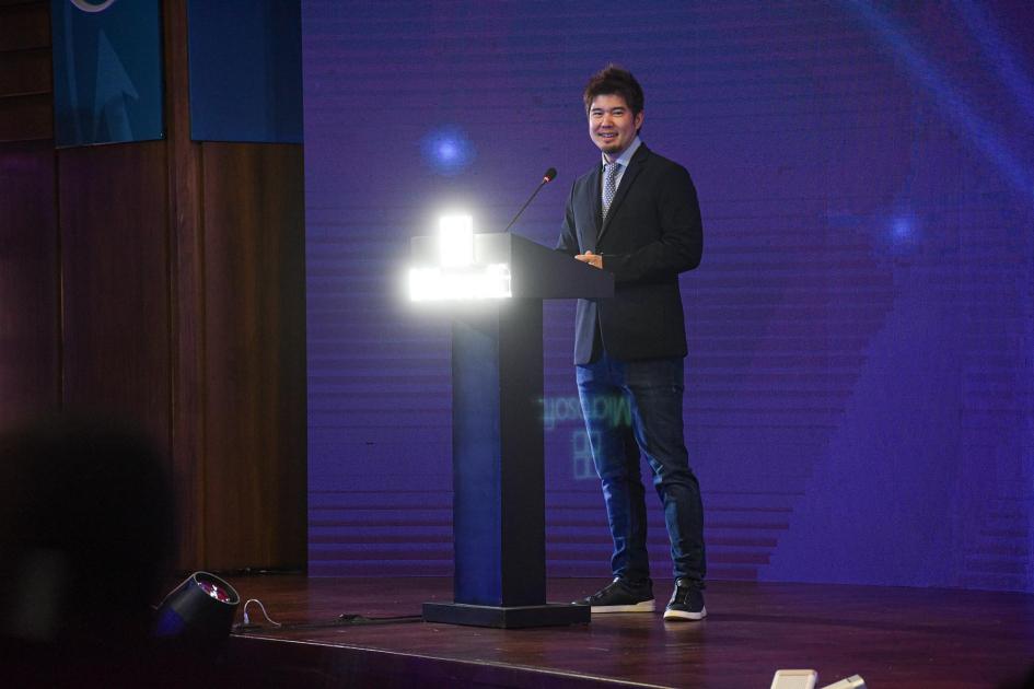 Jun Yamasaki, líder de Microsoft AI Co-Innovation Labs Yamasaki habla desde un atril; detrás, pantalla violeta