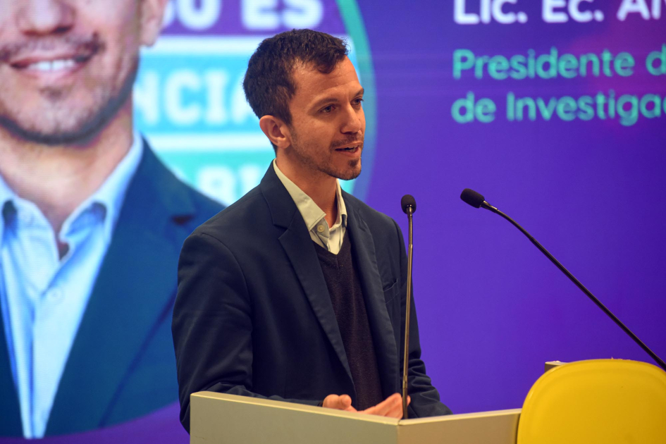 Brunini habla desde un atril; detrás, fragmento de pantalla con su foto y nombre