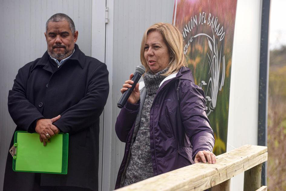 Profesora Mariela del Bono Del Bono habla con un micrófono; detrás, maestro de ceremonias y banner