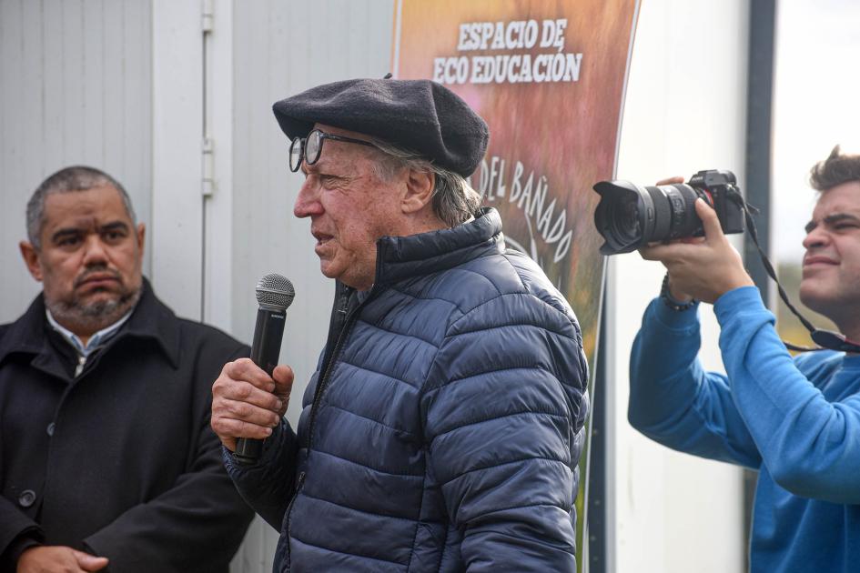 Presidente de la Asociación Bañados de Carrasco, Roberto Garese Garese habla con un micrófono; detrás, fotógrafo, maestro de ceremonias y banner