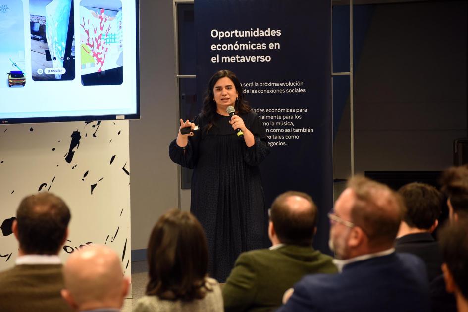 Representante de The Electric Factory explicó en qué consistirá la tecnología de la capacitación Una mujer habla con un micrófono ante los asistentes; a su lado hay una pantalla; detrás, banners