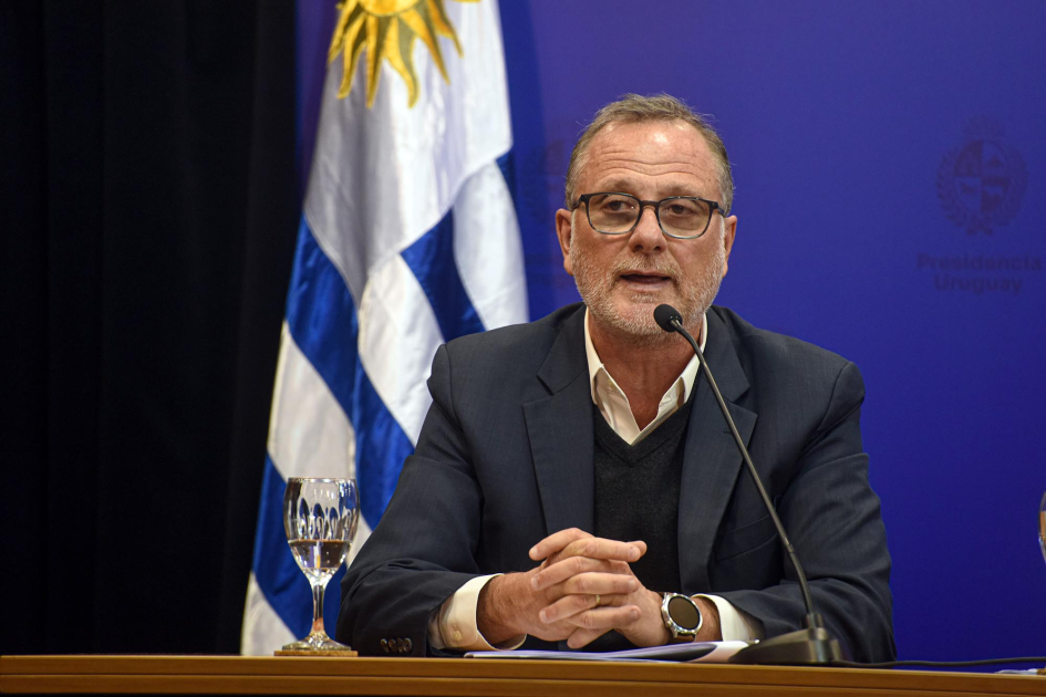 Gili habla, sentado; detrás, bandera uruguaya