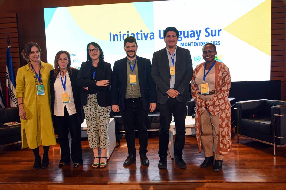 Panel 4 de la primera jornada del encuentro Moderadora y cinco panelistas, de pie; detrás, pantalla