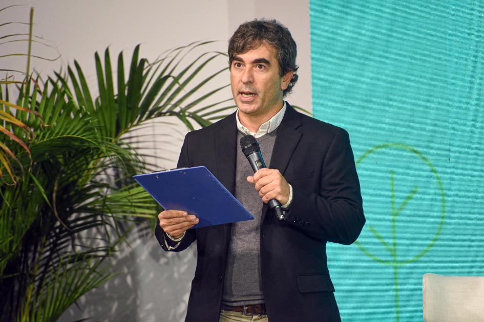 Martín Aguirre, representante del diario El País Aguirre habla con un micrófono y una carpeta en la mano; detrás, planta