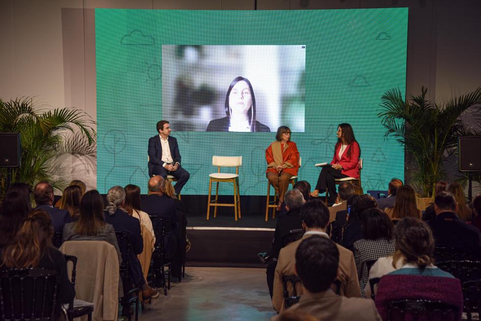 Primer panel de intercambio del evento Tres mujeres sentadas en sillas, frente al público; detrás, otra mujer en una pantalla (vía Zoom)