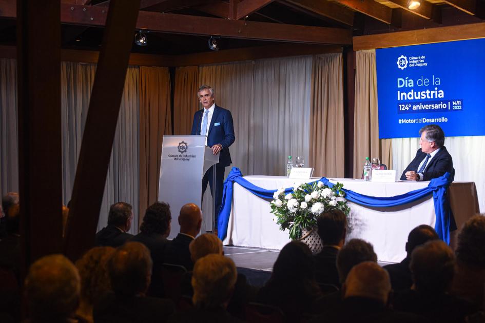 Presidente de la CIU, Alfredo Antía Antía habla desde un atril; en una mesa con mantel blanco y azul, lo observa Paganini