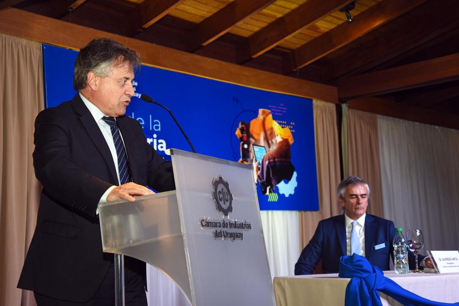 Ministro Omar Paganini expone en el Día de la Industria Paganini habla desde un atril; a su lado, en una mesa, está Antía; detrás, un banner de la fecha