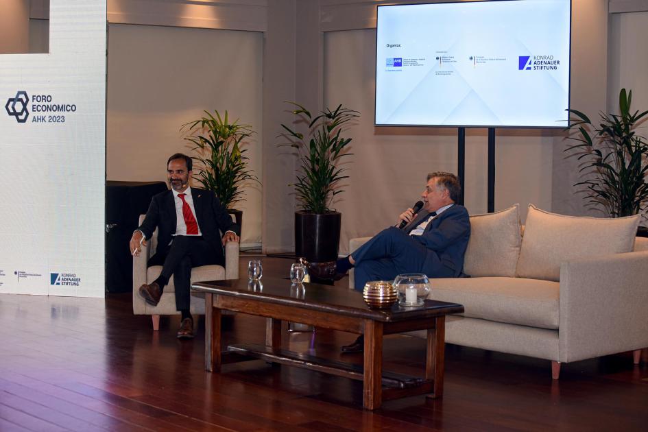 Paganini expuso en la nueva edición del Foro Económico AHK Living en el que dialogan Paganini y otro hombre; detrás hay una pantalla con imagen del evento